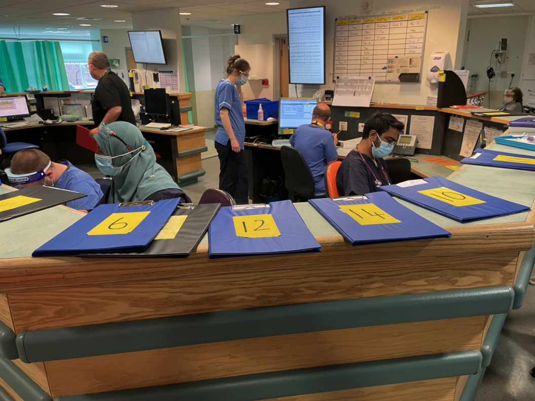 A series of numbered clipboards resting on top of a reception desk, with several masked NHS staff working in the background.