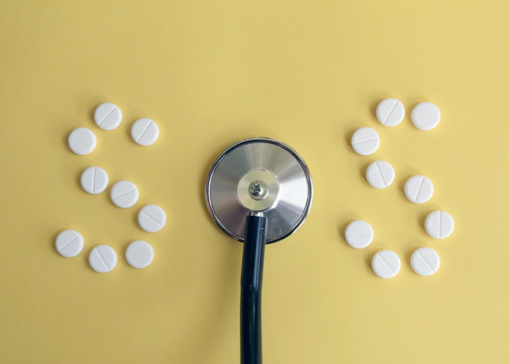 White, circular pills are arranged on a muted yellow background to spell out two 'S' shapes. The circular end of a stethoscope is in the middle of the two 'S' shapes, making the image read SOS.